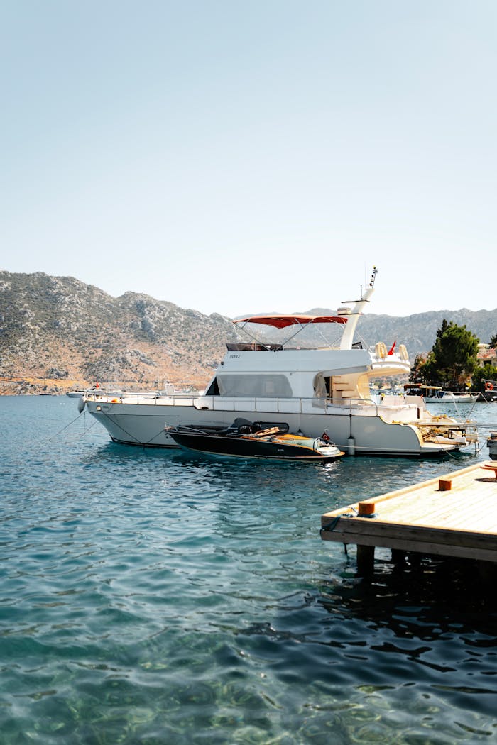 A luxury yacht docked at a serene harbor with clear blue water and scenic mountain backdrop.